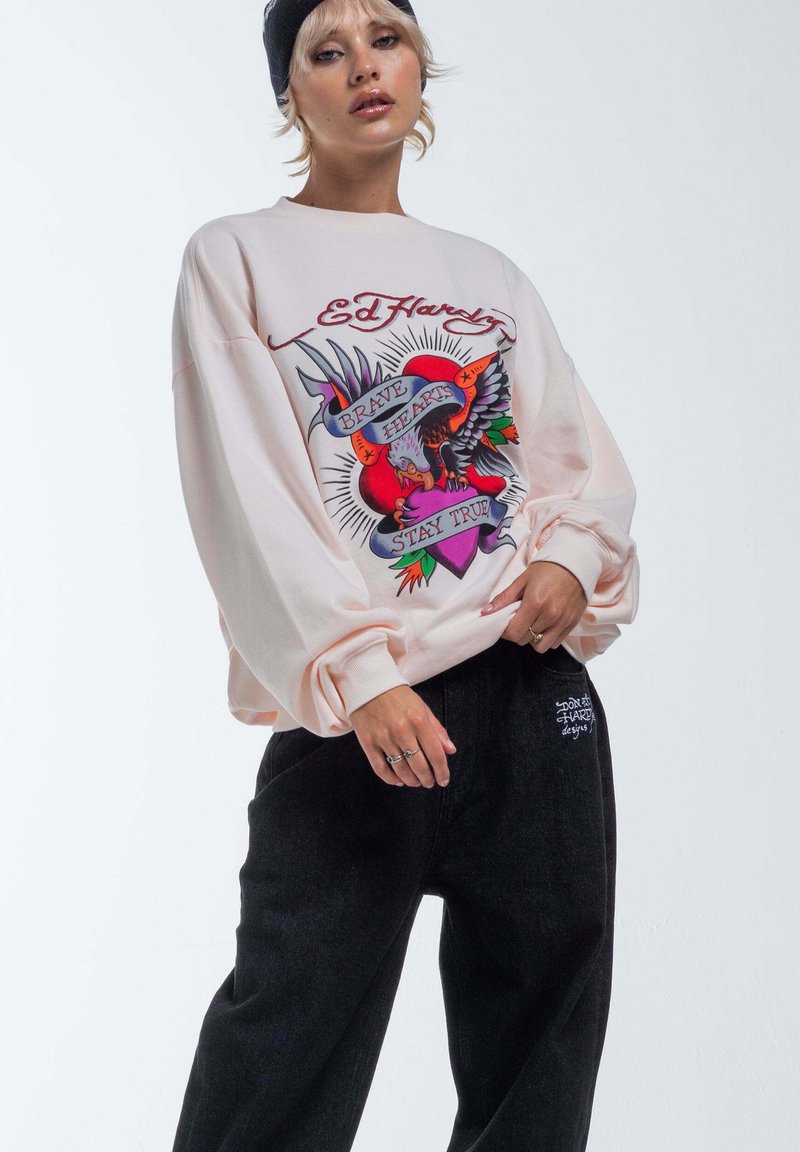 Felpa oversized rosa chiaro con un design grafico colorato che presenta un cuore e un'aquila, abbinata a pantaloni neri larghi.