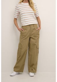 Camisa de manga corta a rayas beige y blanca combinada con pantalones cargo de pierna ancha en color caqui que cuentan con bolsillos. El modelo lleva zapatillas blancas.