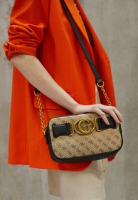 Borsa a tracolla marrone con motivo a fantasia, rifiniture dorate e dettagli neri, tenuta da una persona con blazer arancione, su sfondo neutro.