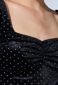Top negro hecho de una tela brillante, con escote cuadrado, detalles fruncidos y un patrón de puntos plateados esparcidos.