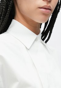 Λευκό πουκάμισο με κουμπιά και κλασικό γιακά, με απαλό υφή και διακριτική γυαλάδα, που δείχνει μέρος του λαιμού και πλεξούδα μαλλιών.