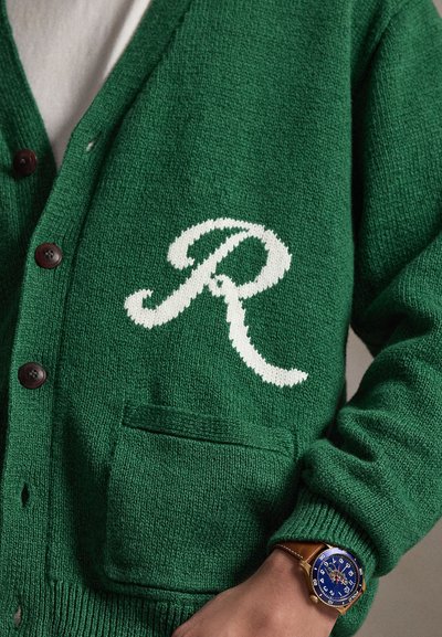 Cardigan verde lavorato a maglia con la lettera bianca "R" sulla tasca indossato da una persona con un orologio da polso blu e oro al polso sinistro.
