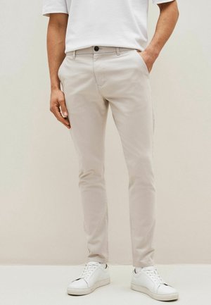 Homme portant un pantalon beige slim-fit, des baskets blanches et un t-shirt blanc, debout avec une main dans la poche devant un fond clair uni.