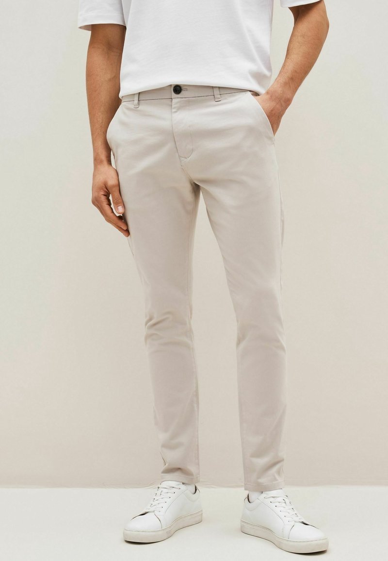 Homme portant un pantalon beige slim-fit, des baskets blanches et un t-shirt blanc, debout avec une main dans la poche devant un fond clair uni.