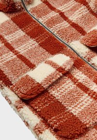 Checkered jacket in orange and cream fleece with a soft texture, featuring a front zipper and two flap pockets.