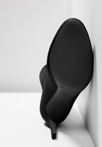 Botte noire à talon haut reposant avec la semelle contre un mur blanc, montrant une semelle en caoutchouc texturée et un talon aiguille fin.