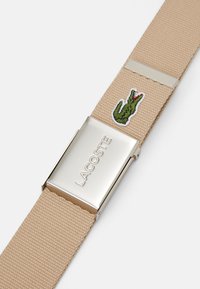 Cinturón tejido beige con una hebilla metálica plateada que presenta el texto grabado "LACOSTE" y un detalle de bordado de cocodrilo verde.