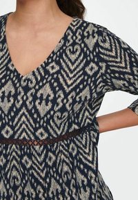 Žena v námořnickém modrém šatu s béžovými geometrickými vzory, s výstřihem do V, tříčtvrtečními rukávy a průhledným detaily v pase.