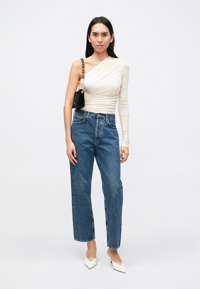 Top assimétrico de mangas compridas em off-white, jeans azuis de perna reta, mala preta com alça de corrente, saltos brancos com bico pontiagudo.