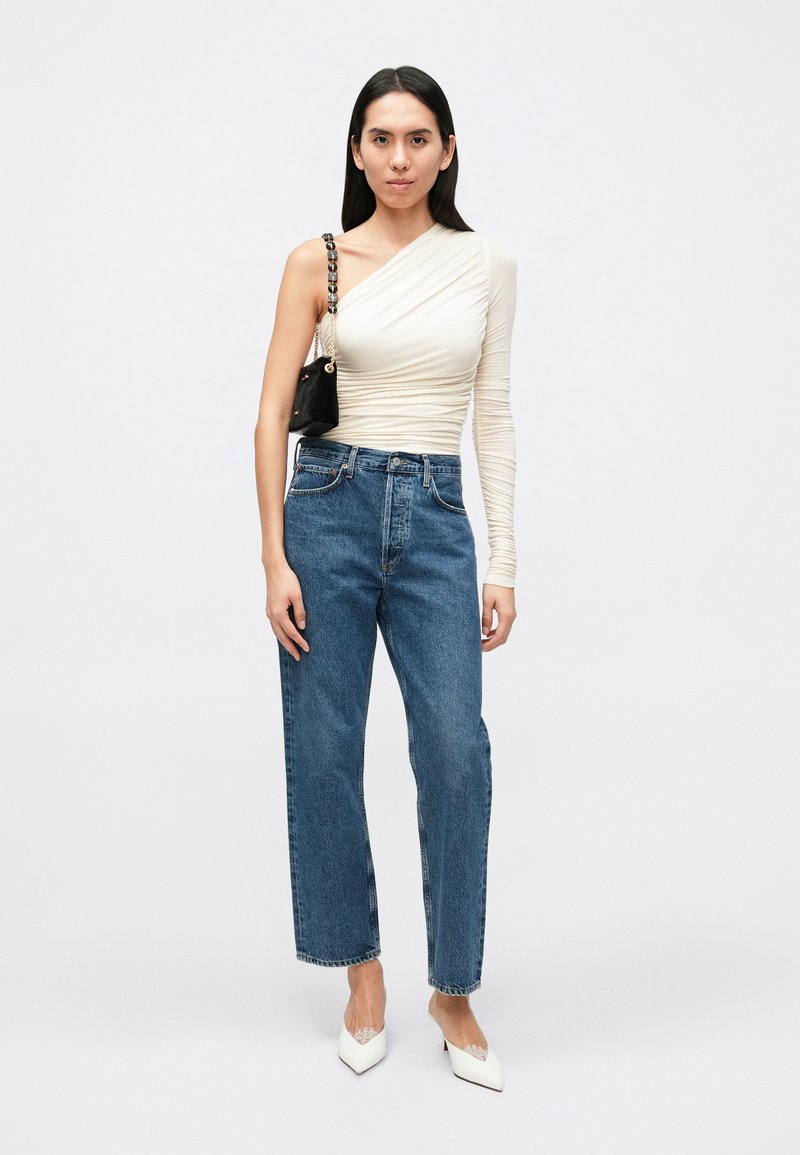 Top asimmetrico a maniche lunghe color avorio, jeans blu a gamba dritta, borsa nera con tracolla a catena, décolleté bianche con punta affilata.