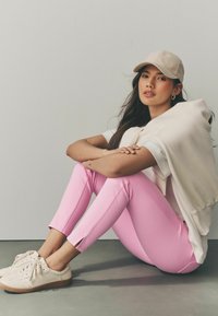 Pantalones ajustados rosas con aberturas laterales, combinados con una sudadera crema caída sobre los hombros y zapatillas beige. Pose casual y relajada.