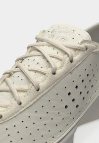 Κοντινό πλάνο μπεζ αθλητικού παπουτσιού Nike Air με διάτρητο πλαϊνό πάνελ και κορδόνια που σταυρώνονται πάνω από τη μαξιλαρωμένη γλώσσα.