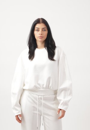 Ung kvinde med langt mørkt hår iført en oversized hvid sweatshirt og matchende hvide bukser, der står foran en ensfarvet hvid baggrund.