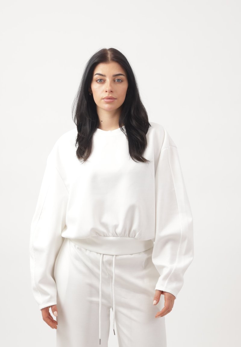 Giovane donna con lunghi capelli scuri che indossa una felpa bianca oversize e pantaloni bianchi coordinati, in piedi davanti a uno sfondo bianco uniforme.