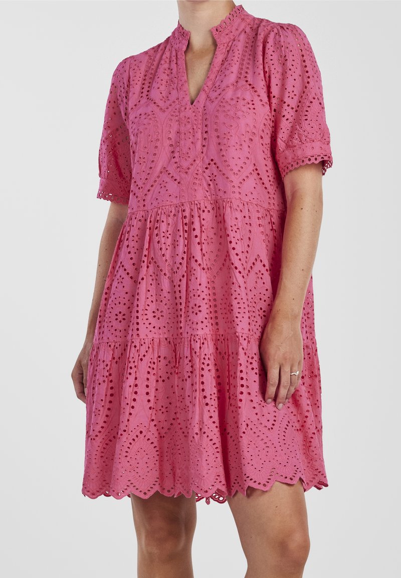 Robe rose à manches courtes avec œillets, ourlet festonné, col en V et motifs floraux brodés portée par une personne debout.