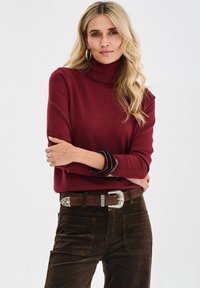 Frau mit langen blonden Haaren in einem burgunderfarbenen Rollkragenpullover, braunen Cordhosen, einem breiten Gürtel mit reich verziertem Schnallen und schwarzen Armbändern.