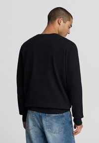 Uomo con i capelli corti che indossa un maglione nero semplice e jeans blu, di spalle contro uno sfondo grigio chiaro uniforme.