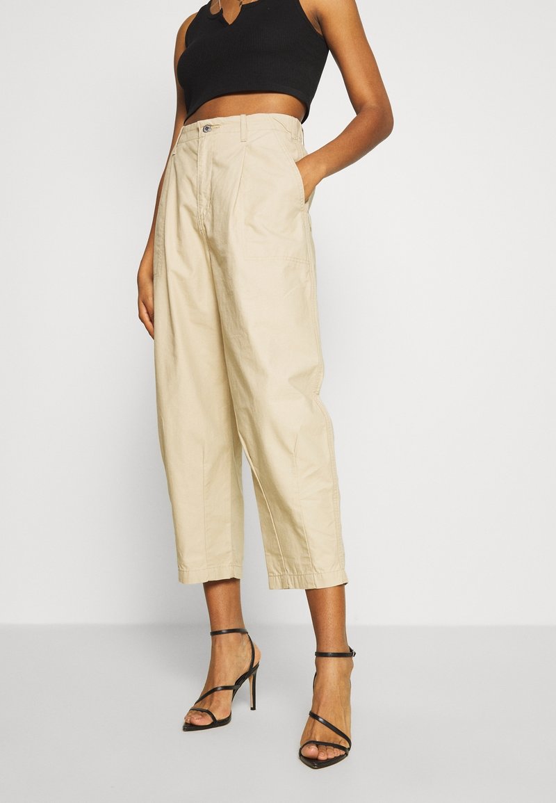Femme portant un pantalon beige large et court, un crop top noir sans manches et des sandales noires à talons hauts à lanières, debout avec une main dans la poche.