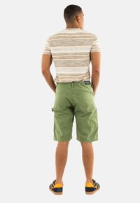 Shorts cargo verts avec poches arrière, associés à une chemise à rayures beige et marron à manches courtes. Le modèle porte des chaussures brunes et jaunes.