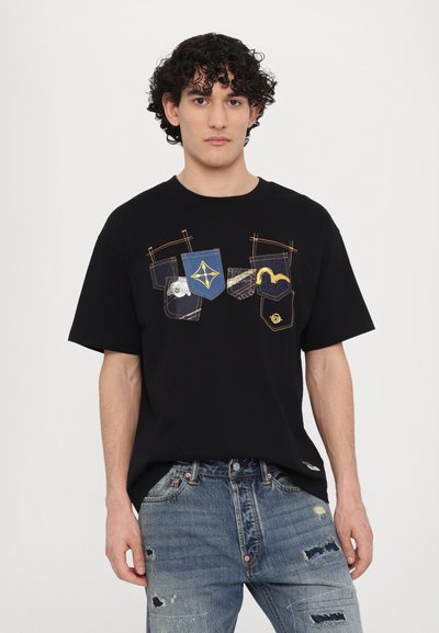 T-shirt en coton noir avec des designs graphiques sur la poche en bleu, jaune et blanc. Manches courtes et coupe décontractée. Porté avec un jean en denim déchiré.