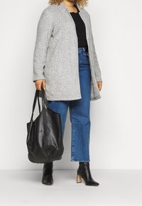 Grauer Wollmantel mit geradem Schnitt, Fronttaschen, über einem schwarzen Pullover getragen. Kombiniert mit schwarzen High Heels und einer schwarzen Lederhandtasche.