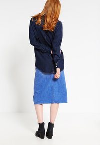 Mörk denimskjorta med lång ärm, rundad nederkant och knappar vid cuffs, i kombination med en ljusblå denimpencilkjol och svarta klackboots.