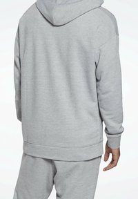 Personne portant un sweat à capuche gris clair texturé et un pantalon assorti, vue de dos sur un fond blanc.