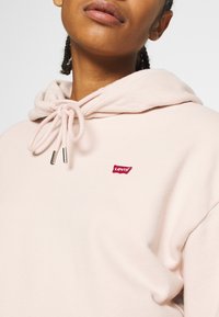 Närbild av en person som bär en ljusrosa Levi's hoodie med dragsnoddar knutna i en rosett och en liten röd Levi's-logotyp på bröstet.