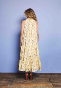 Žena s kučeravými vlasmi má na sebe žlté kvetované šaty s ramienkami a hnedé sandále na podpätku, stojí na drevenej podlahe pred svetlofialovou stenou.