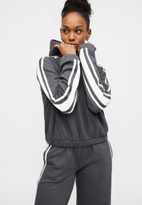 Γκρι φούτερ με κουκούλα και αντίστοιχο παντελόνι φόρμας με λευκές ρίγες στα μανίκια και στις πλευρές. Μαλακή υφή, ελαστικές μανσέτες και ζώνη στη μέση.