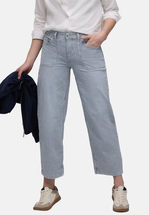 Person iført hvid skjorte og blå-hvidstribede korte bukser med hvide sneakers, der holder en navy jakke i venstre hånd.