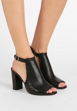 Sandalias de tacón abiertas en la punta, de cuero negro con un acabado texturizado, costados abiertos y una correa en el tobillo con hebilla dorada. Diseño de tacón cuadrado.