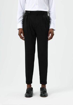 Black pleated pants with a tailored fit, featuring a waistband button and side pockets, styled with rolled cuffs and paired with black shoes.