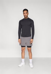 Μαύρο μακρυμάνικο τοπ και γκρι αθλητικό σορτς φορεμένα πάνω από μαύρο σορτς συμπίεσης, συνδυασμένα με λευκά αθλητικά παπούτσια και κάλτσες μέχρι τον αστράγαλο.