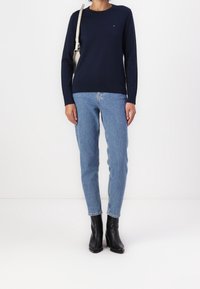 Granatowy sweter z okrągłym dekoltem w zestawieniu z jasnoniebieskimi jeansami oraz czarnymi botkami na obcasie do kostki. Na swetrze widoczne jest małe logo.