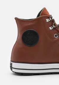 Baskets montantes en cuir marron avec semelle en caoutchouc noire, œillets argentés et lacets verts. Présente un patch logo circulaire noir sur le côté.