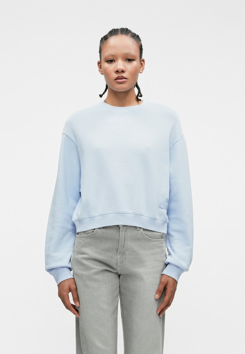 Ανοιχτό μπλε κοντό φανελάκι με μακρύ μανίκι, στρογγυλή λαιμόκοψη και λεπτομέρεια με τσέπη. Συνοδεύεται από γκρι τζιν ίσιου γραμμής.