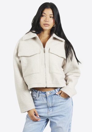 Vrouw met lang donker haar, gekleed in een beige cropped jas met grote zakken en een rits, en lichtblauwe jeans met hoge taille, staand met één hand in de zak.