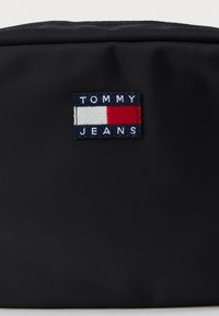 Fekete nylon tasak, elöl egy fehér, piros és kék téglalap alakú logóval, amelyen az "TOMMY JEANS" felirat található. Sima felület. Cipzáras záródás.