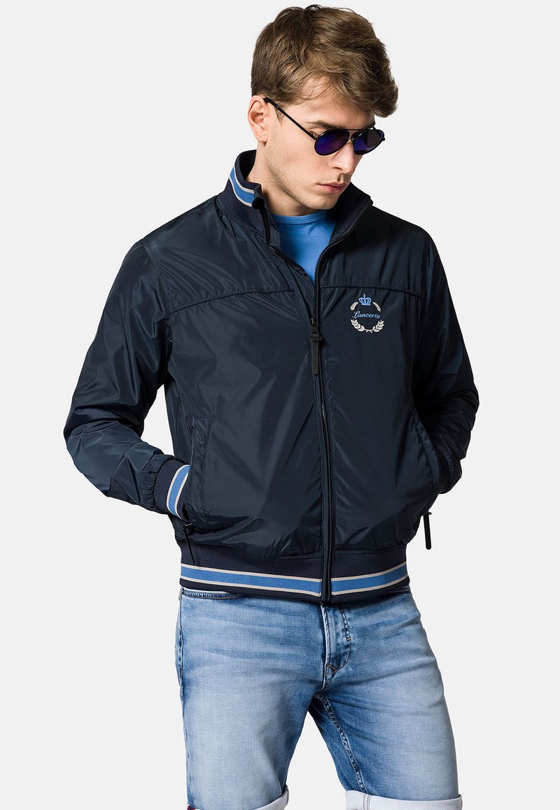 Granatowa bomberka z błyszczącego materiału, z prążkowanymi ściągaczami przy mankietach i u dołu, zapinana na zamek, z haftowanym logo na klatce piersiowej.