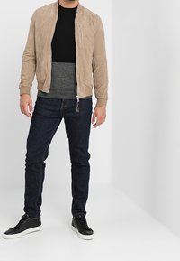 Beige Wildleder-Bombers jacket mit Reißverschluss-Brusttasche, schwarzem Rundhalsausschnitt-Pullover, grauem Mittelteil, dunkelblauen Jeans und schwarzen Sneakers.