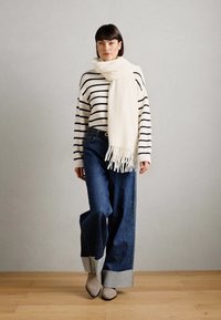 Pull rayé blanc, écharpe crème avec des franges, jeans en denim sombre à coupe large et bottines taupe. Design simple, texture douce, look décontracté.
