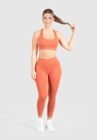 Oranžová sportovní podprsenka a odpovídající vysoce pasové legíny vyrobené z elastického materiálu. Modelka má na sobě bílé sportovní boty s jednoduchým designem.