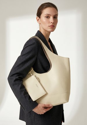Vrouw in een donker colbert houdt een grote beige gestructureerde leren shopper vast met bijpassend klein tasje tegen een lichte achtergrond.