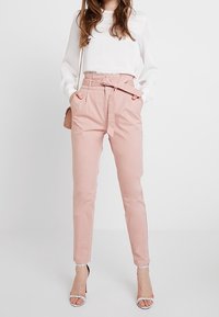 Pantalon taille haute rose pâle en mélange de coton, avec un détail de ceinture, des jambes fuselées et des poches latérales, associé à un haut blanc.