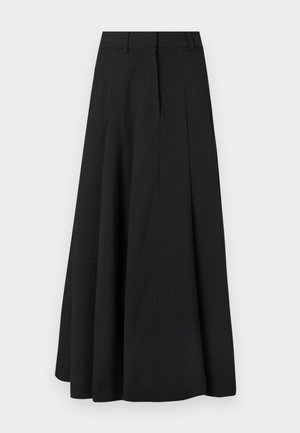 Femme portant un cardigan côtelé noir et une jupe plissée noire, tenant un sac à main beige froncé, debout sur un sol gris avec un fond de mur uni.
