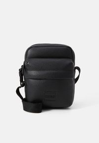 Černá kožená crossbody taška s texturovaným povrchem, zipovým uzávěrem, nastavitelným popruhem a přední logo nášivkou. Kompaktní obdélníkový tvar.