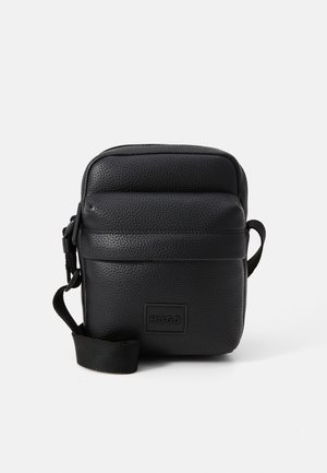 Sac bandoulière en cuir noir avec surface texturée, fermeture éclair, sangle réglable et un patch logo à l'avant. Forme rectangulaire compacte.