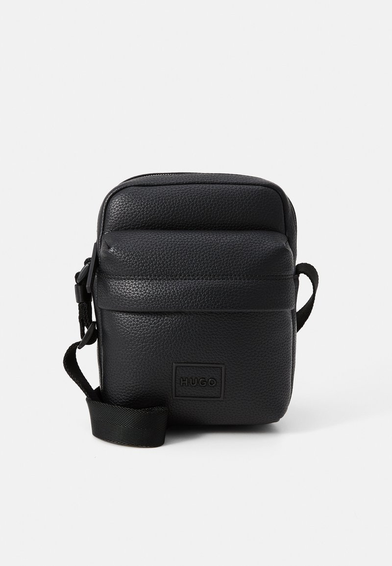 Černá kožená crossbody taška s texturovaným povrchem, zipovým uzávěrem, nastavitelným popruhem a přední logo nášivkou. Kompaktní obdélníkový tvar.