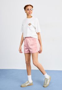 Weißes T-Shirt mit einem kleinen, mehrfarbigen Grafikdesign, kombiniert mit rosa Satin-Shorts mit weißen Streifen und grauen Sneakers mit weißen Akzenten.